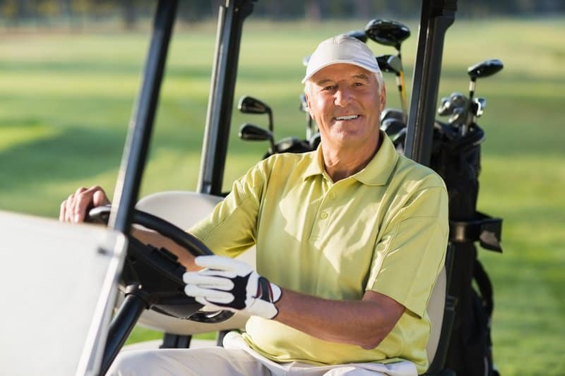 A senior golfer riding and driving a golf cart with a golf bag on a golf course